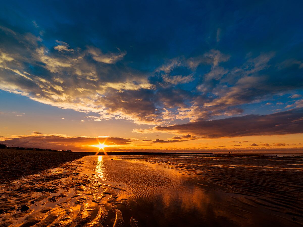 Dramatischer Sonnenuntergang am Strand mit spiegelndem Wasser, goldenem Licht und blauem Wolkenhimmel. Zwei Personen in der Ferne.