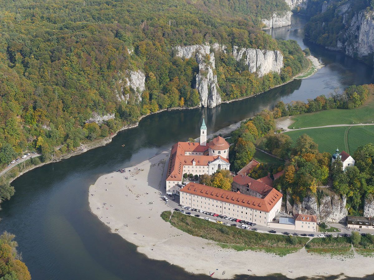 Luftaufnahme von Kloster Weltenburg an der Donau, umgeben von herbstlichen Wäldern, weißen Felsen und einem Sandstrand mit Menschen.
