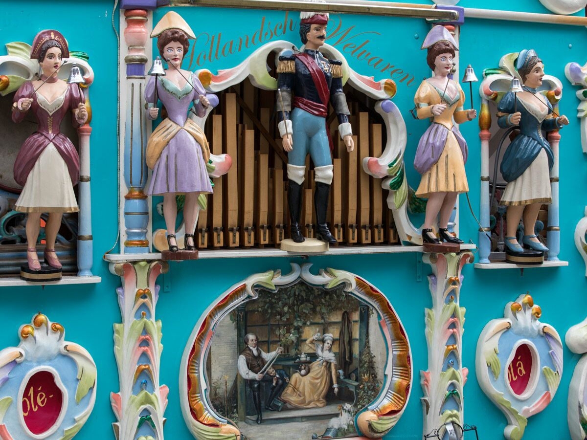 Historische Kirmesorgel mit animierten Figuren (Dirigent, Frauen mit Glocken) vor blauer Wand.