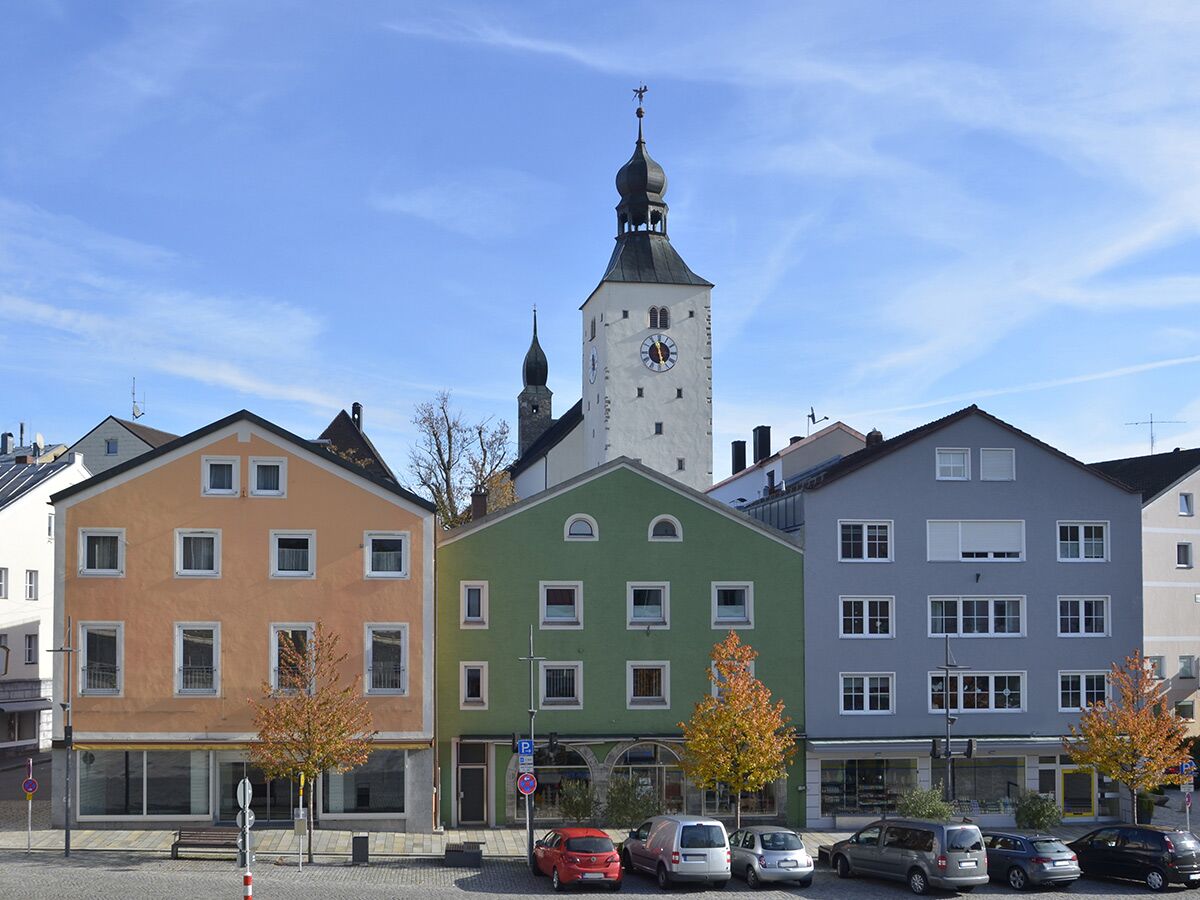 Straße mit bunten Häusern in einer Altstadt, vorne ein orangefarbenes, grünes und graues Gebäude, im Hintergrund ein hoher Kirchturm.