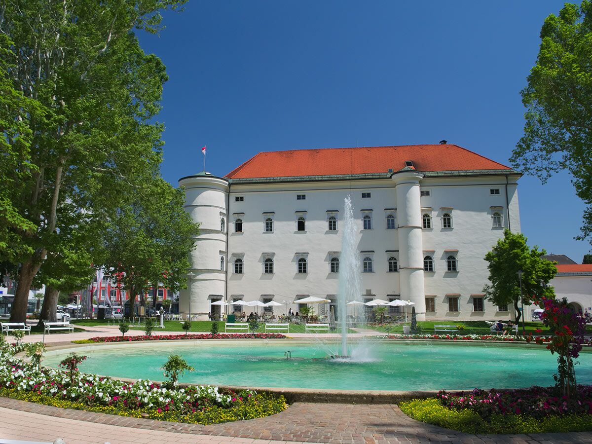 Schloss Hartberg, Österreich: historisches Gebäude mit rotem Dach, großer Brunnen mit Wasserspiel, Blumen und Bäumen im sonnigen Park.