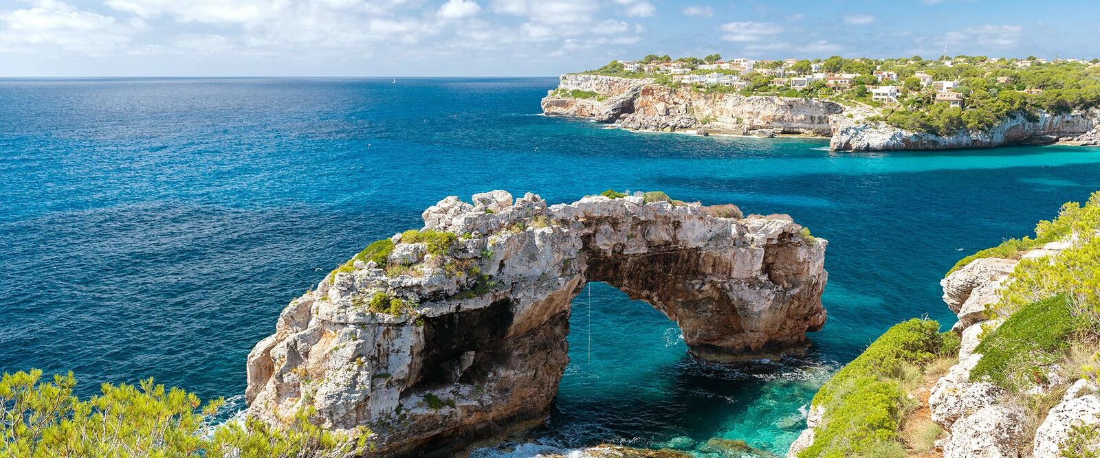 Atemberaubender Blick auf den natürlichen Felsbogen Es Pontàs auf Mallorca mit türkisfarbenem Mittelmeer und Küstenort.