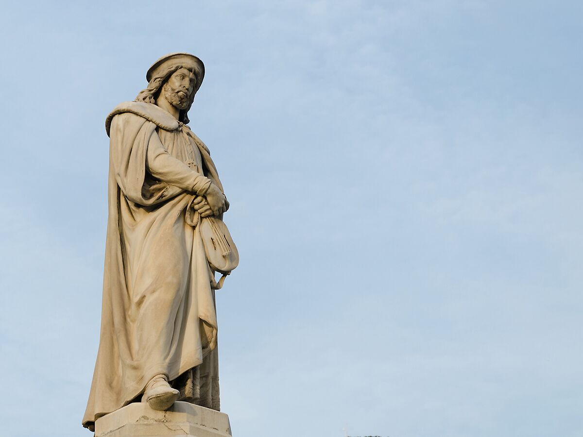 Stein-Statue eines bärtigen Mannes in historischer Kleidung mit Hut und Umhang, der eine Laute hält, vor blauem Himmel.