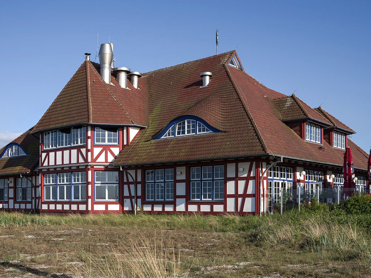 Traditionelles Strandhaus Restaurant im Fachwerkstil mit rotem Ziegeldach, weißen Wänden, blauen Fensterrahmen und Terrasse unter blauem Himmel.