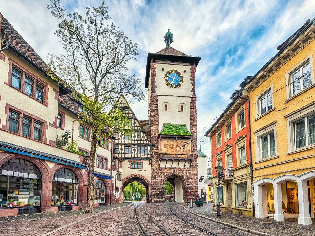 Historisches Schwabentor in Freiburg mit Uhr, Torbogen, Kopfsteinpflaster, Straßenbahnschienen und Fachwerkhäusern unter blauem Himmel.