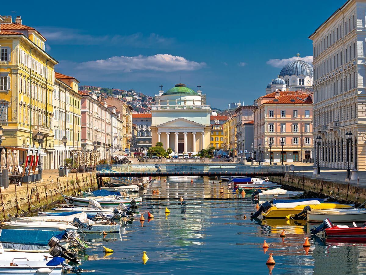 Malerischer Kanal in Triest, Italien, mit bunten Gebäuden, historischen Kirchen und vielen kleinen Booten bei Sonnenschein.