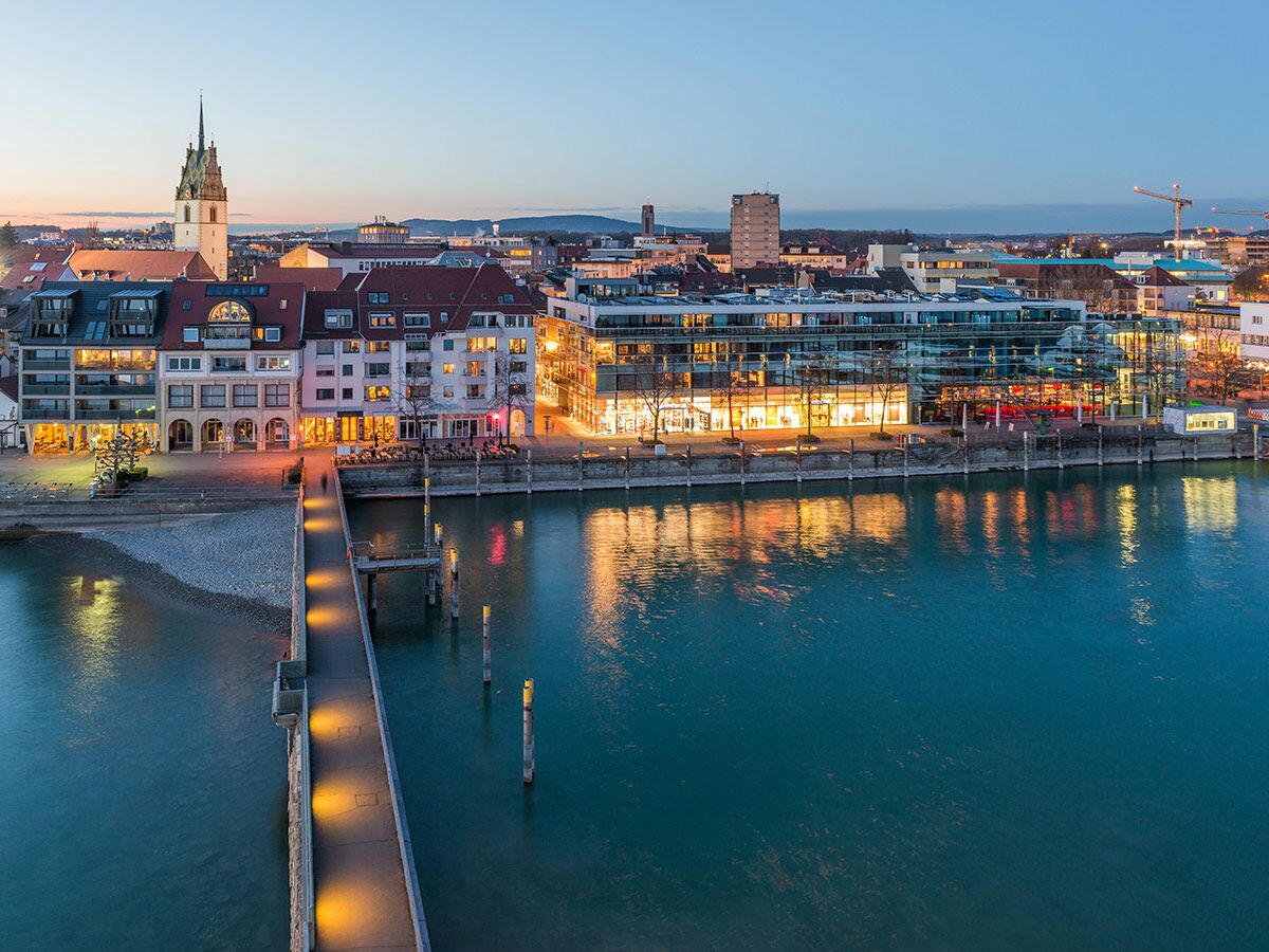 Abendansicht der Konstanzer Seepromenade mit historischer Architektur, modernem Gebäude, beleuchtetem Steg und Bodensee bei Dämmerung.