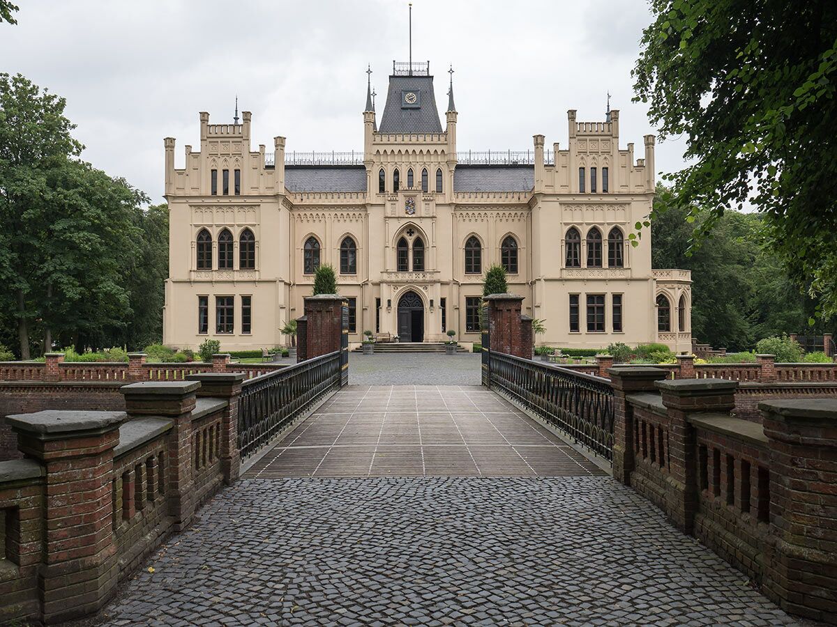 Neogotisches Schloss Brincke mit hellem Sandstein, zentralem Turm, dunklem Dach, von Brücke und Park umgeben.