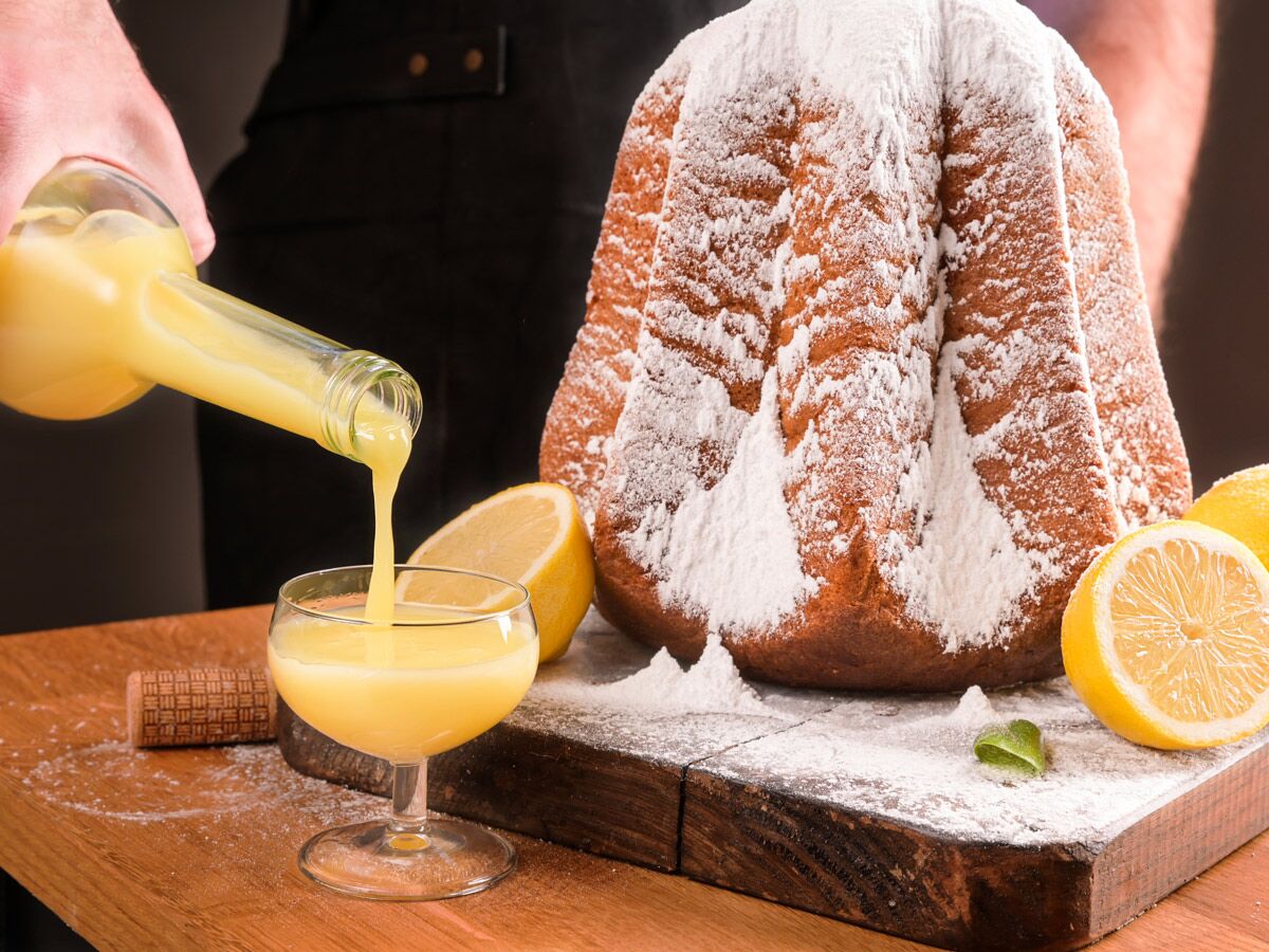 Ein Pandoro-Kuchen mit Puderzucker und frischen Zitronen, während Limoncello in ein Glas auf einem Holzbrett gegossen wird.