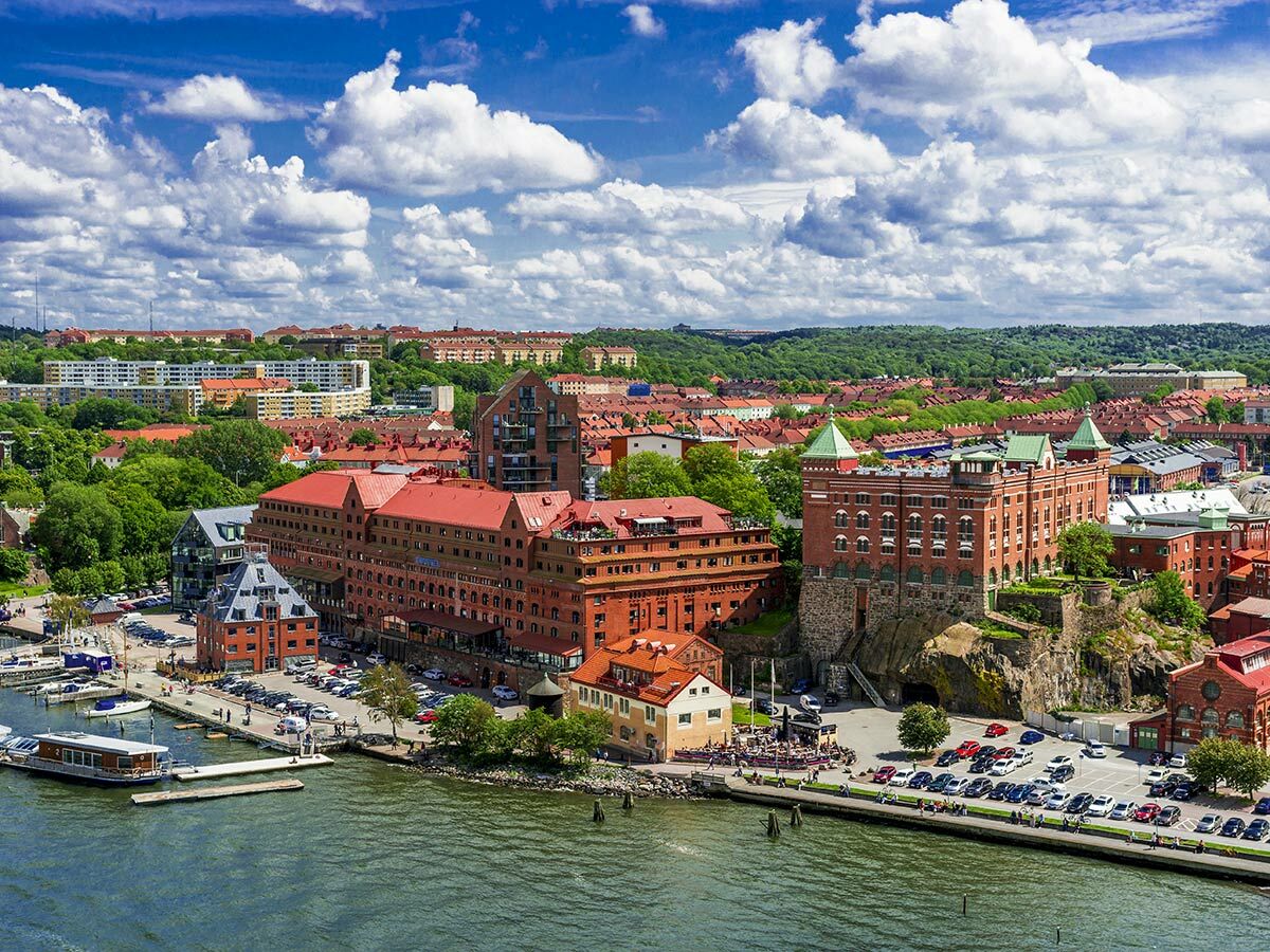 Luftaufnahme der Göteborger Uferpromenade mit roten Ziegeldächern, Booten im Hafen und Stadtsilhouette unter blauem Himmel.