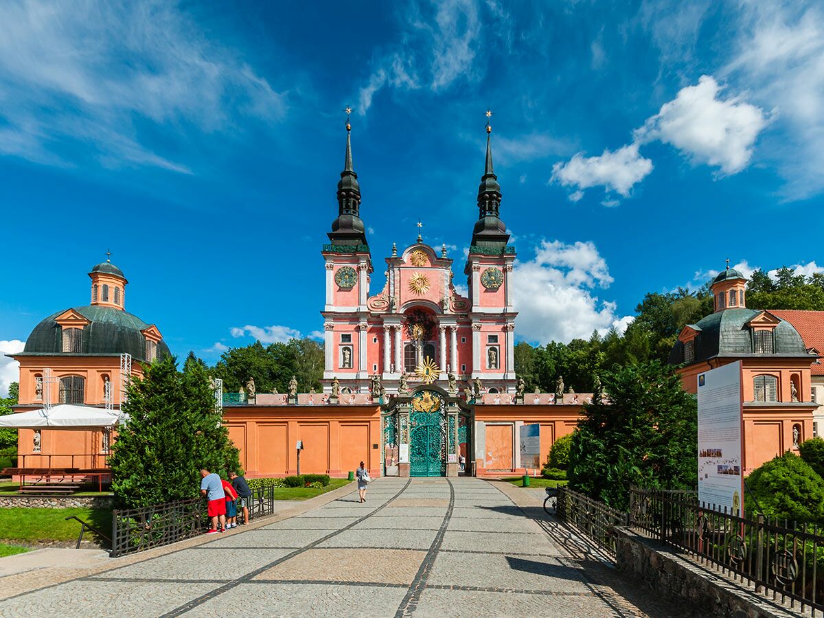 Prachtvolle rosa Barockkirche mit zwei Türmen und orangefarbenen Gebäuden der Abtei Krzeszów in Polen unter blauem Himmel mit Besuchern.