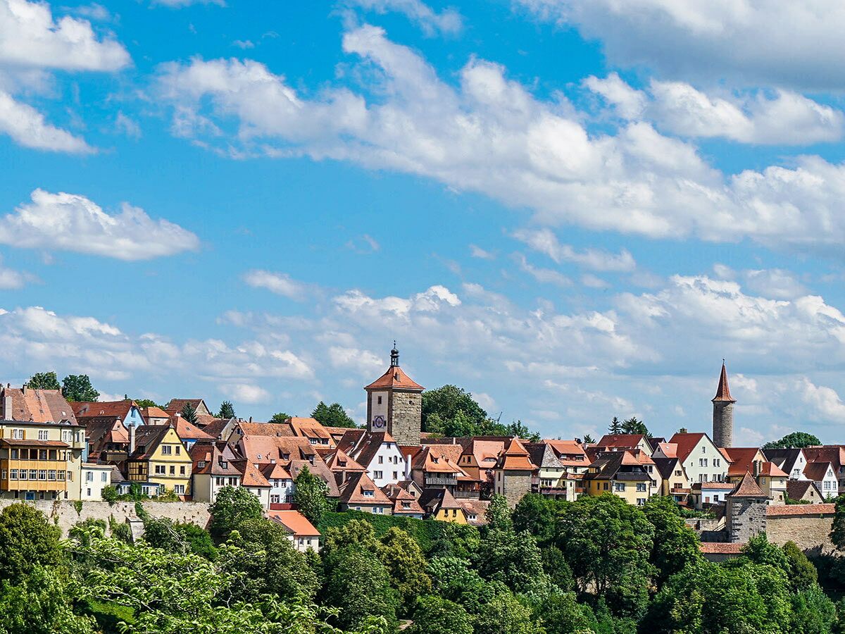 Historische Stadtansicht von Rothenburg ob der Tauber mit Fachwerkhäusern, roten Dächern, Türmen und grünem Vordergrund unter blauem Himmel.