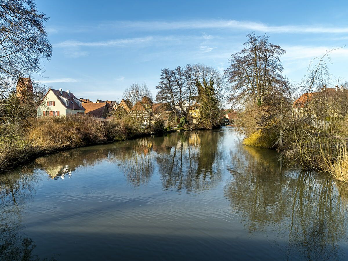 Malerischer Flusslauf mit historischen Häusern und Bäumen an beiden Ufern, die sich im ruhigen Wasser spiegeln. Blauer Himmel.