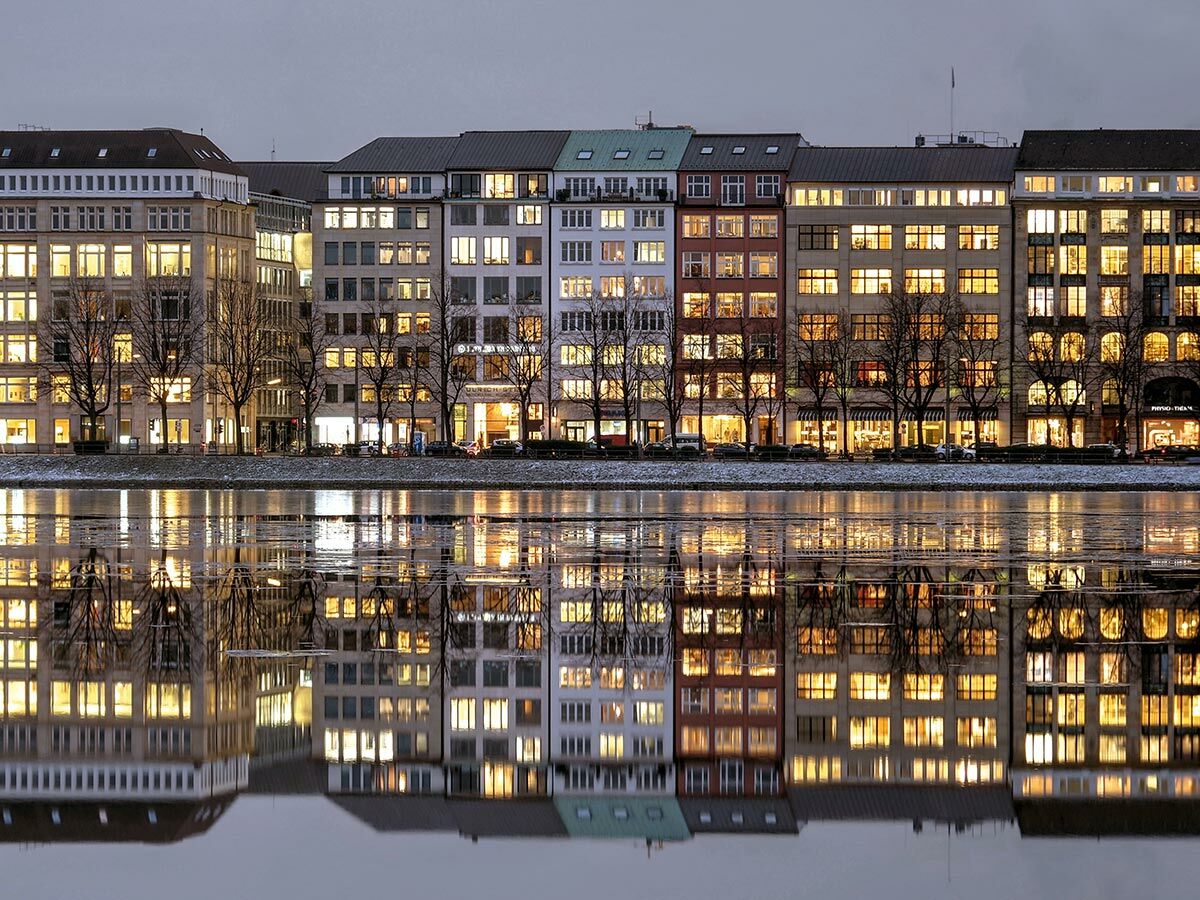 Beleuchtete Stadtgebäude spiegeln sich bei Winterabend im teilweise zugefrorenen Wasser. Warme Lichter erhellen die Fassaden.