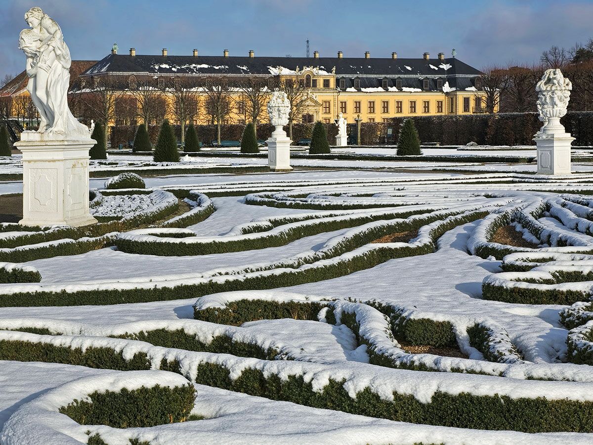 Barocker Schlossgarten im Winter mit Schnee auf kunstvollen Hecken, weißen Statuen und gelbem Palastgebäude im Hintergrund.