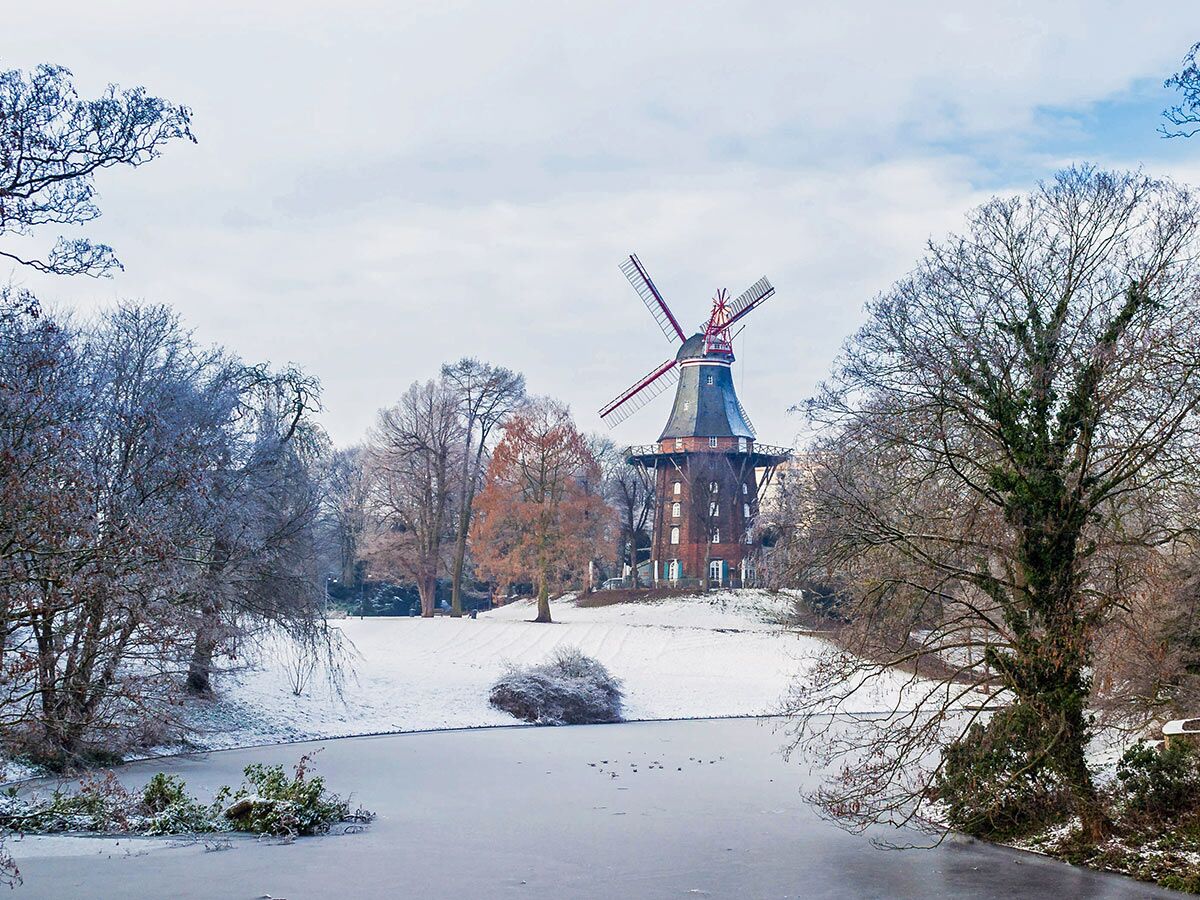 Historische Mühle am Wall in Bremen im Winter, umgeben von Schnee, Bäumen und einem gefrorenen Teich.