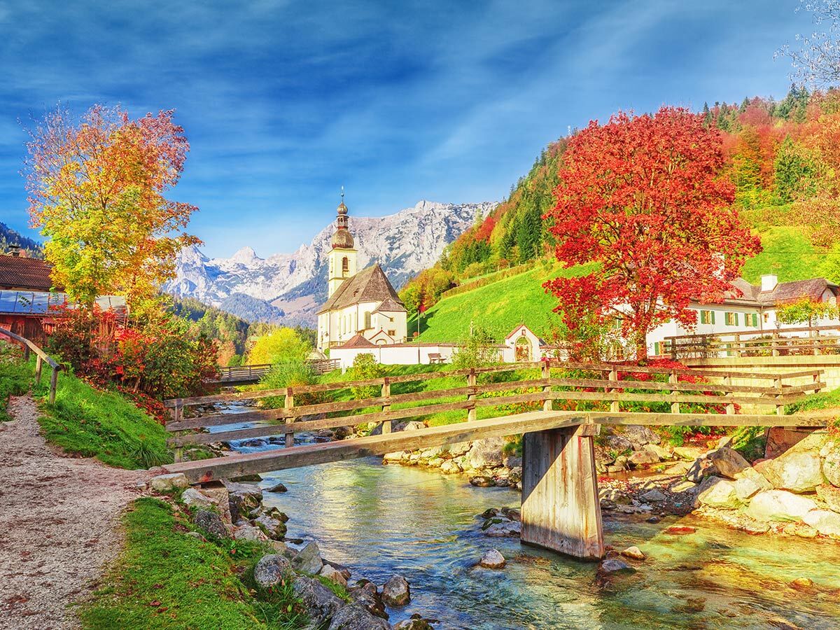 Malerisches bayerisches Alpendorf im Herbst mit Kirche, Fluss, Brücke und bunten Bäumen vor schneebedeckten Bergen.