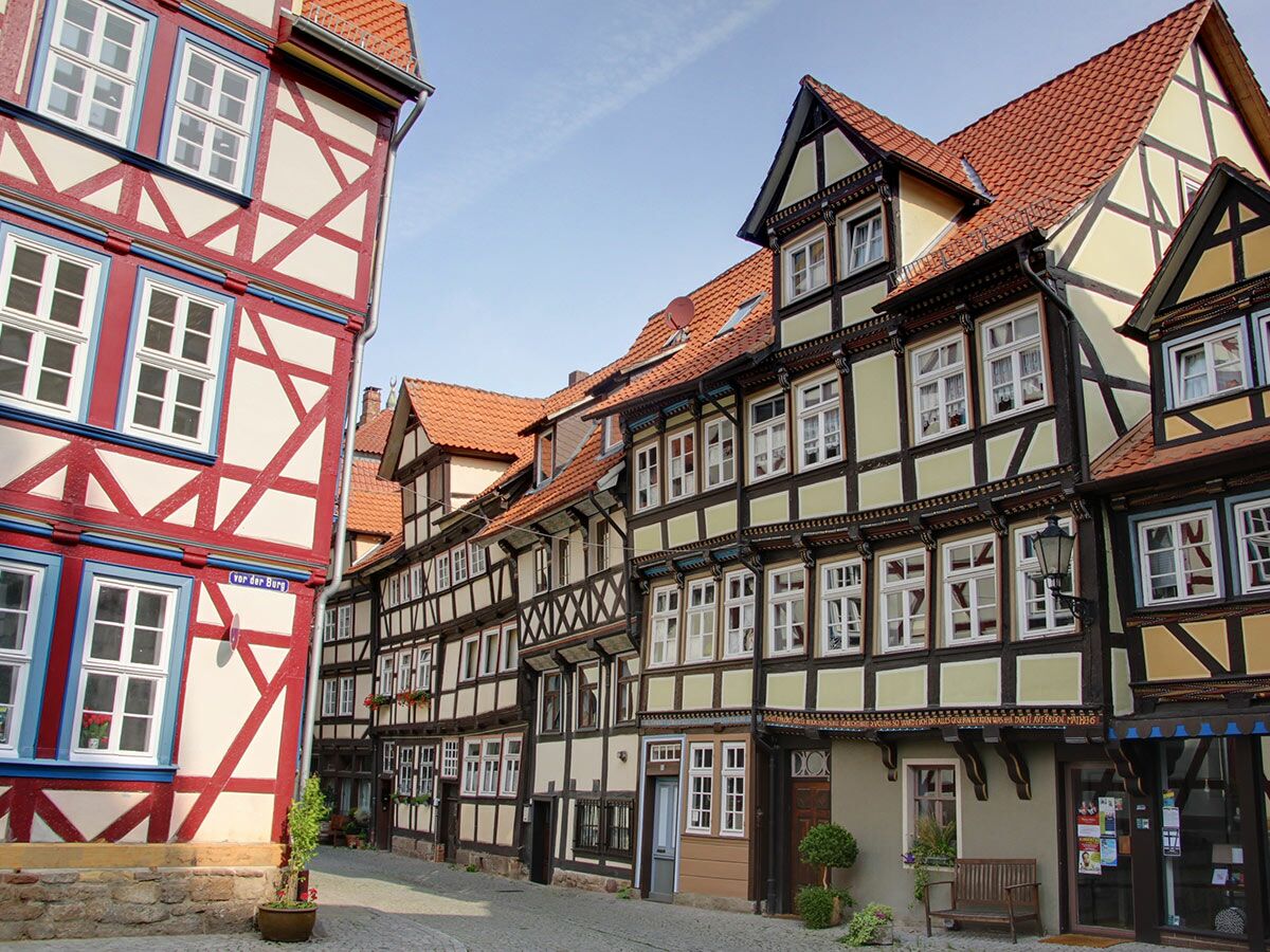 Historische Fachwerkhäuser in einer Gasse mit Kopfsteinpflaster, blauer Himmel. Links rotes Haus, rechts gelbe und braune Fassaden.