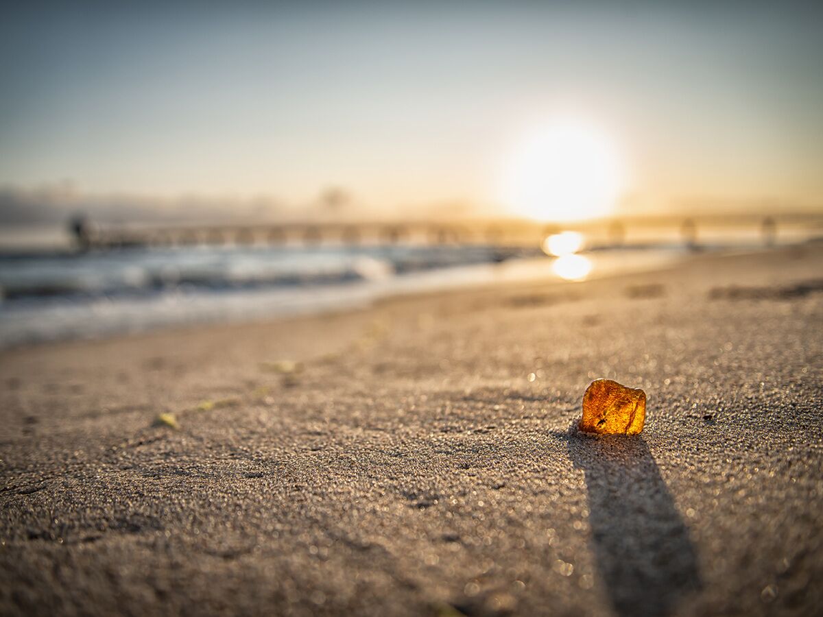 Bernstein am Strand im goldenen Sonnenlicht bei Sonnenuntergang oder -aufgang, Meer mit Wellen und Pier im Hintergrund.