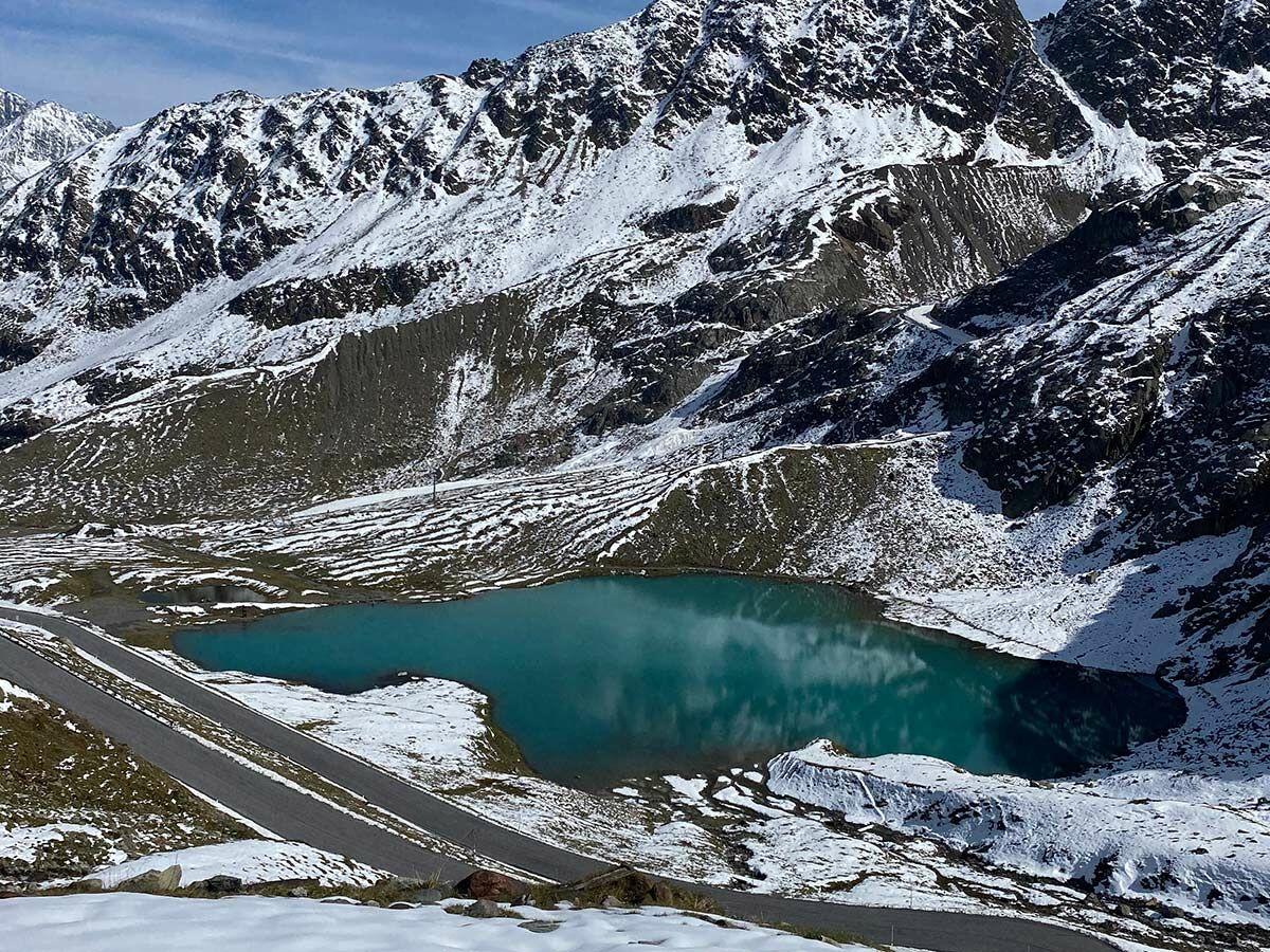 Malerische Alpenlandschaft mit schneebedeckten Gipfeln, einem türkisfarbenen Bergsee und einer Serpentinenstraße.