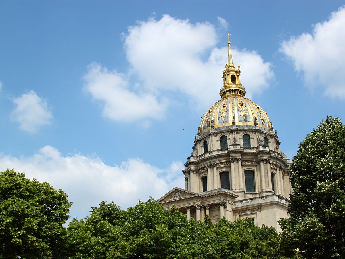 Goldene Kuppel des Dôme des Invalides in Paris unter blauem Himmel mit weißen Wolken und grünen Bäumen an einem sonnigen Tag.