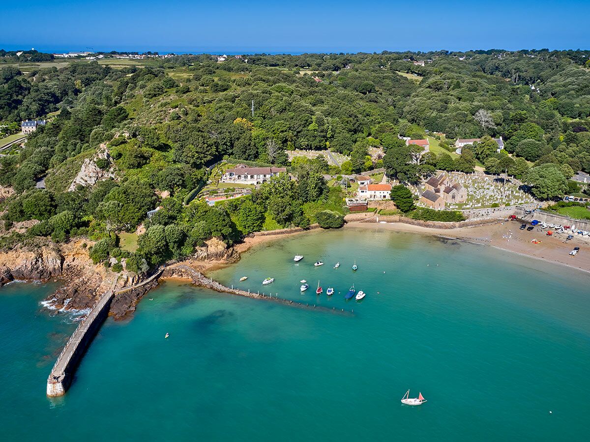 Luftaufnahme einer idyllischen Bucht mit türkisfarbenem Wasser, Strand, Booten, einer Kirche, einem Pier und grünen Hügeln.