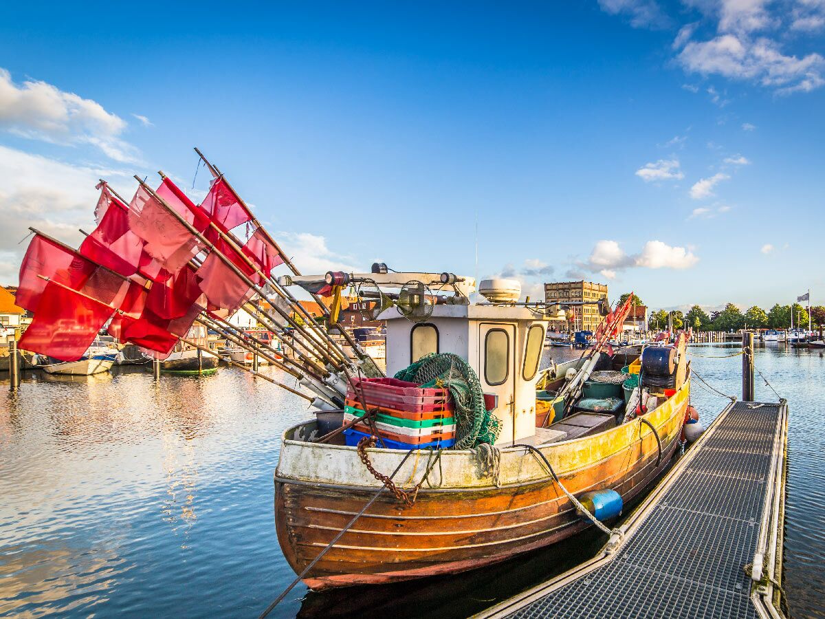 Traditionelles Fischerboot mit roten Fangflaggen und Netzen liegt im sonnigen Hafen am Pier vor Gebäuden.