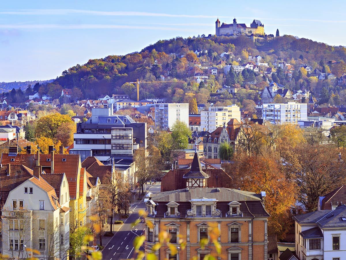 Panorama einer historischen deutschen Stadt mit imposanter Burg auf einem bewaldeten Hügel und bunten Herbstbäumen unter blauem Himmel.