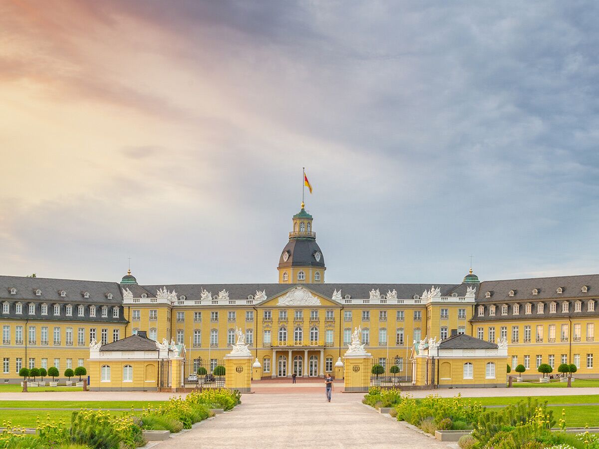 Das Schloss Karlsruhe, ein gelber Barockpalast mit Kuppel, deutscher Flagge und grünen Gärten unter Wolken.