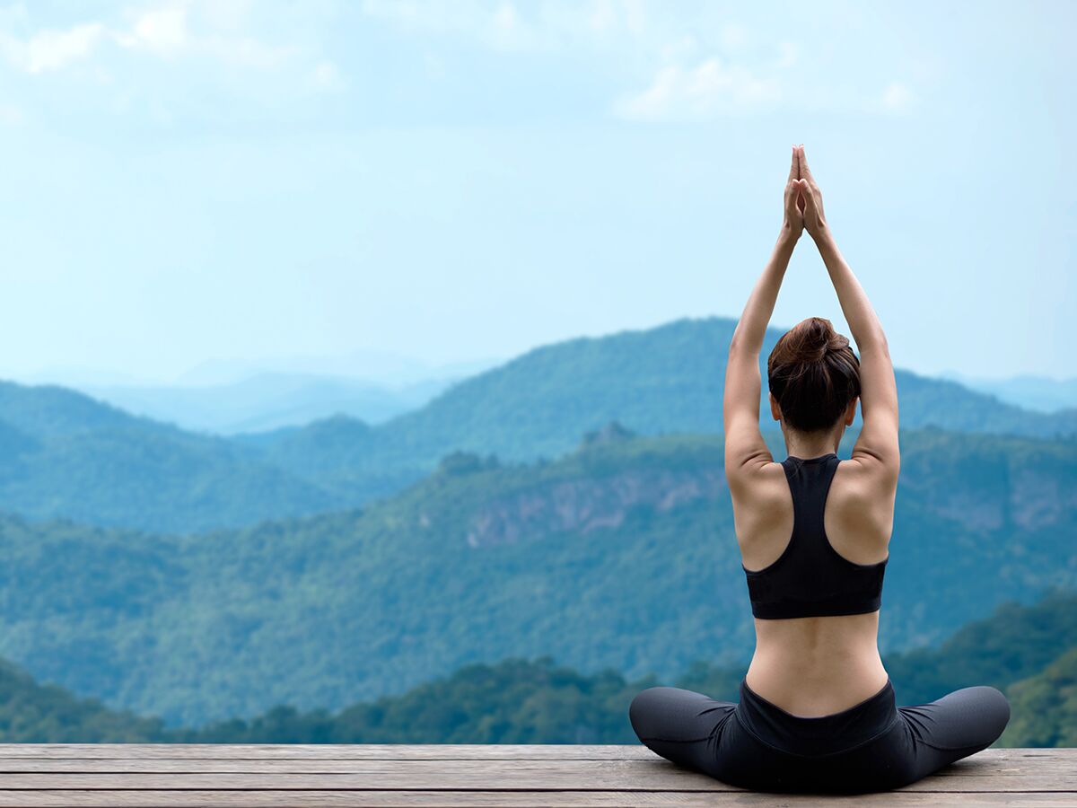 Frau praktiziert Yoga im Freien auf einer Holzplattform, mit erhobenen Armen und Blick auf eine weite Berglandschaft.