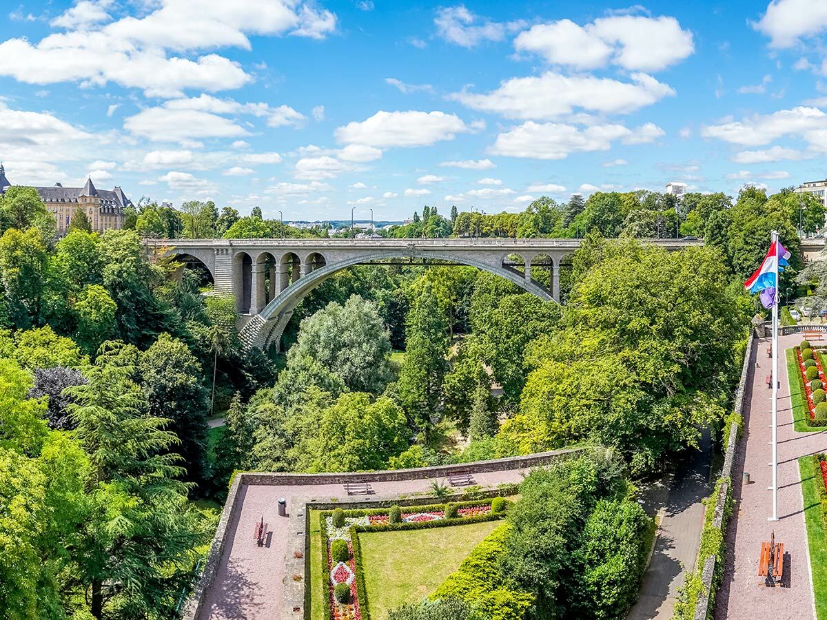 Panoramablick auf Luxemburg-Stadt mit der Adolphe-Brücke über das grüne Pétrusse-Tal und einen lebhaften Park.