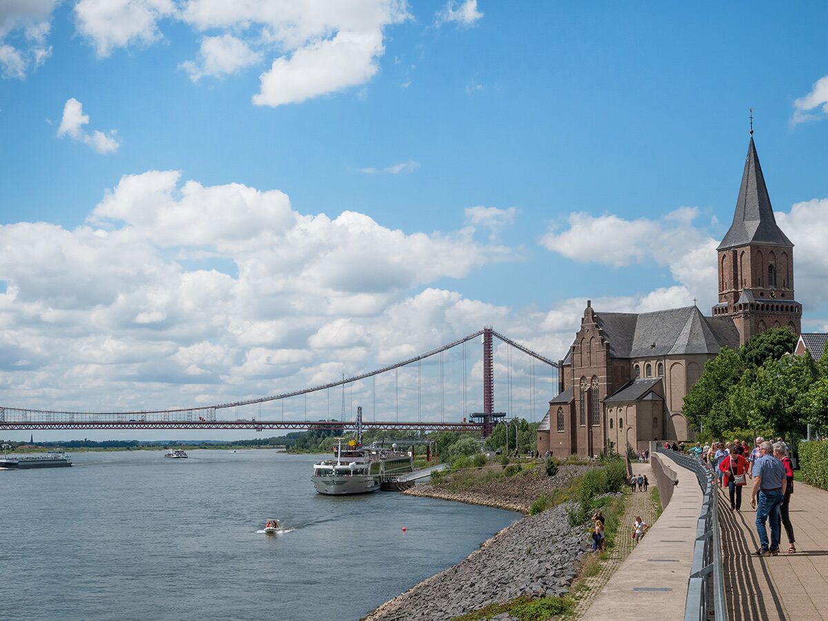 Panoramablick auf Flusslandschaft mit historischer Backsteinkirche, großer Hängebrücke, Booten und Menschen am Ufer unter blauem Himmel.