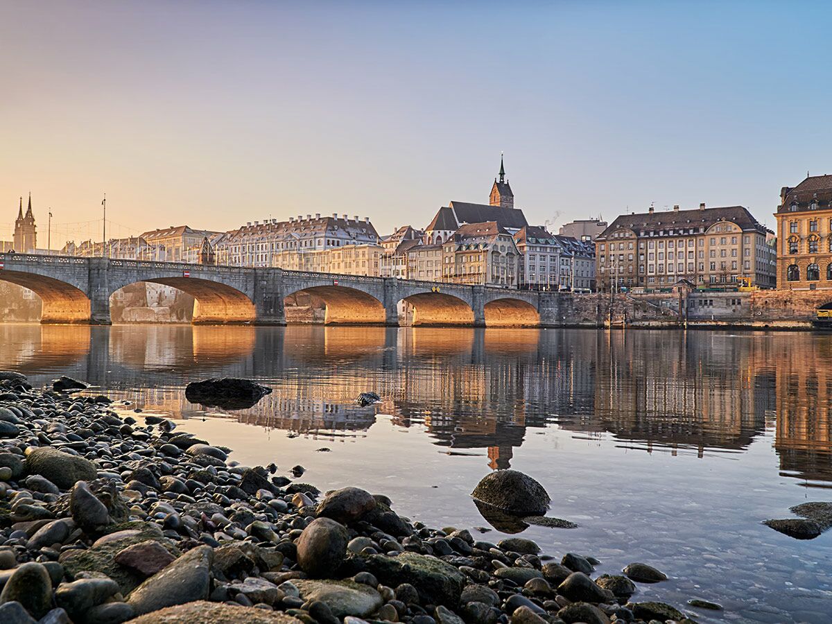 Sonnenaufgang über der Mittleren Brücke Basel mit malerischem Stadtbild am Rhein und Spiegelungen im Wasser.