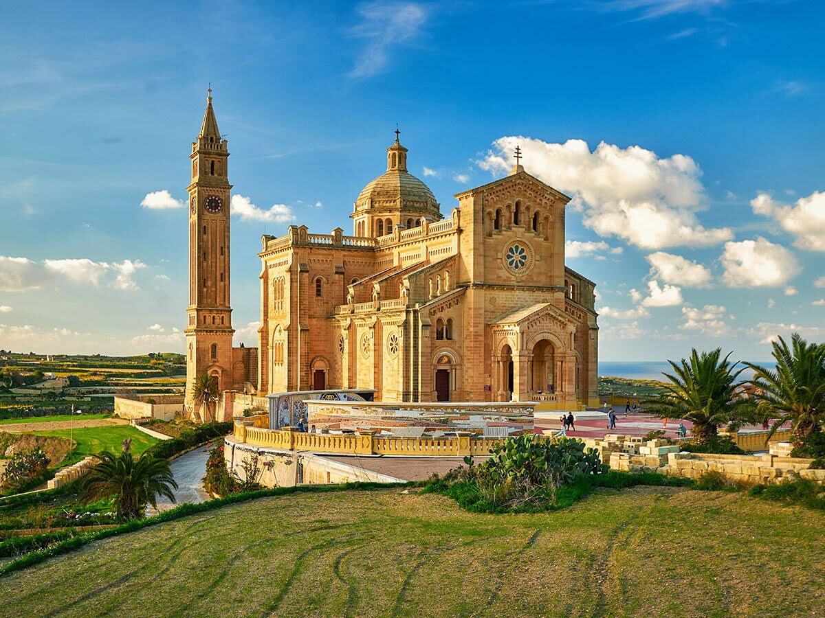 Das Ta' Pinu Heiligtum auf Gozo, Malta, mit hohem Uhrturm, Kuppel, grünen Feldern und dem Mittelmeer.