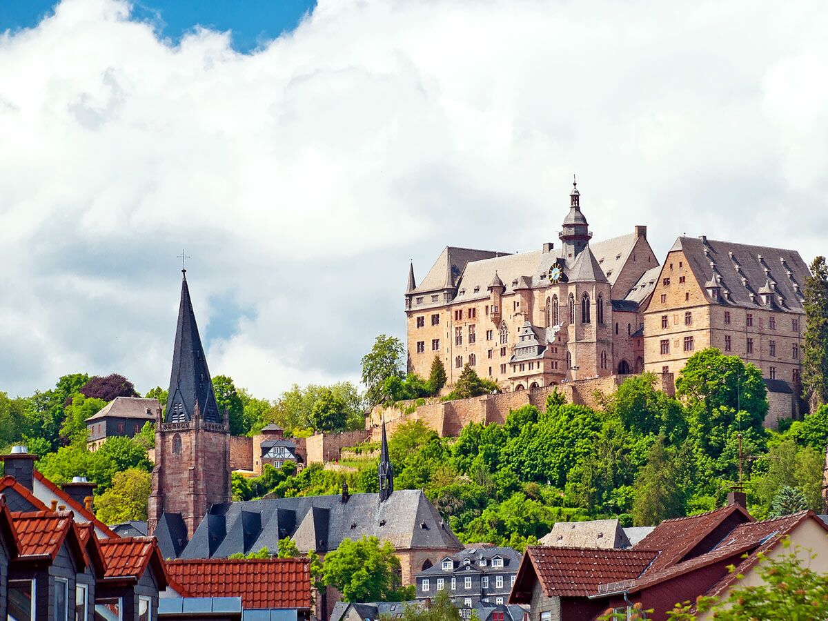 Historisches Marburg mit Schloss, Elisabethkirche und Fachwerkhäusern auf grünen Hügeln unter bewölktem Himmel.