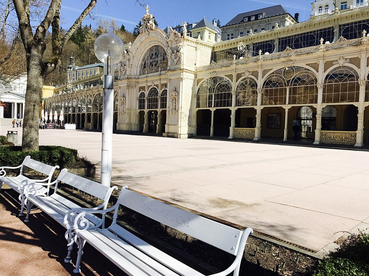 Historische Kolonnade in Mariánské Lázně mit weißen Bänken und hellem Platz an einem sonnigen Tag.