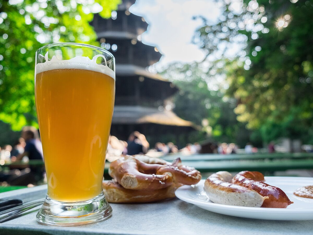 Kühles Weißbier, Brezeln und Wurst auf einem Tisch im sonnigen Biergarten. Im Hintergrund Bäume und ein Pagodenturm.