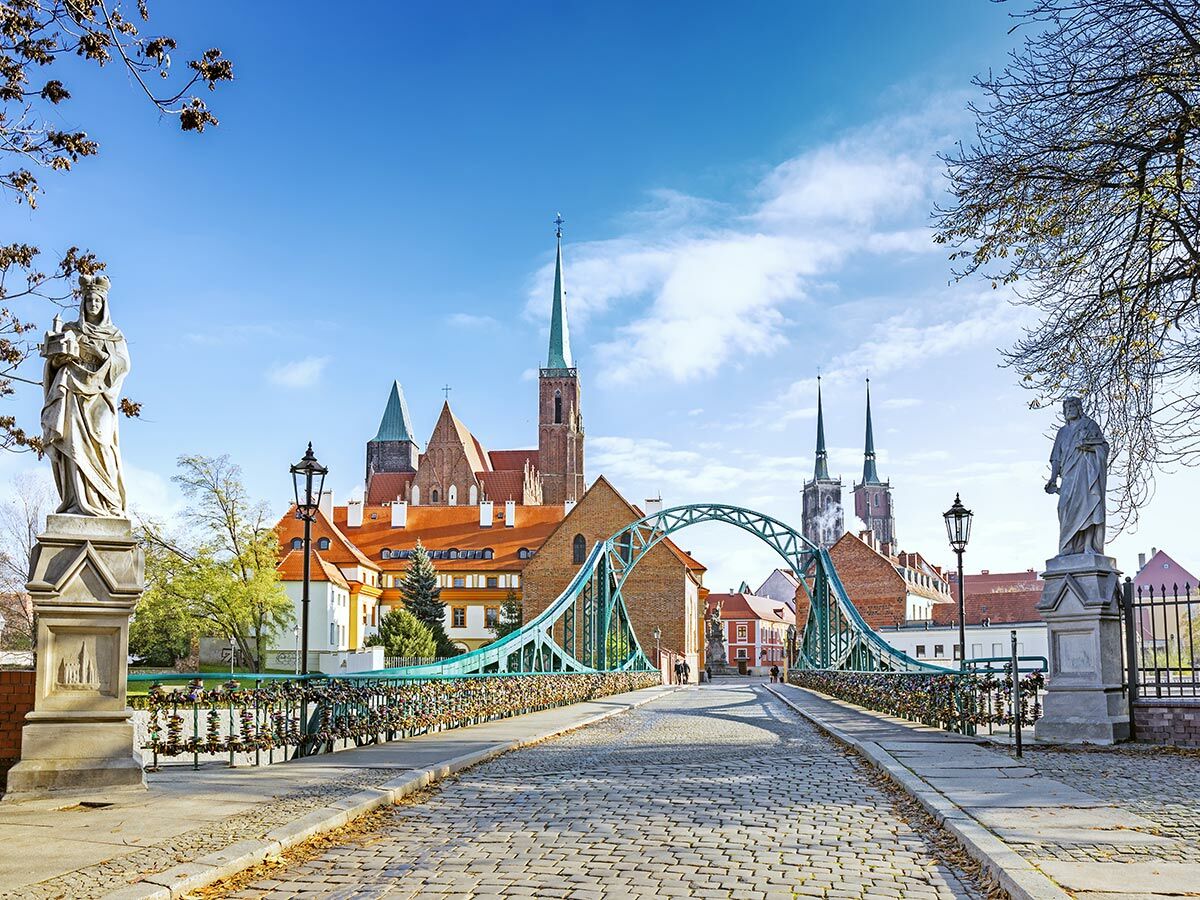 Blick auf die Tumski Brücke in Wrocław mit Liebesschlössern, Statuen und der Kathedrale im Hintergrund bei blauem Himmel.