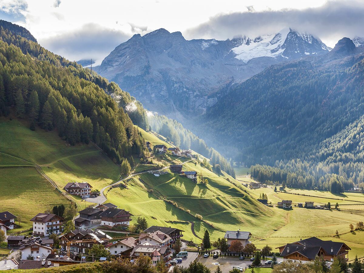 Malerisches alpines Bergdorf in einem grünen Tal mit Wäldern und schneebedeckten Bergen, beleuchtet von Sonnenstrahlen.