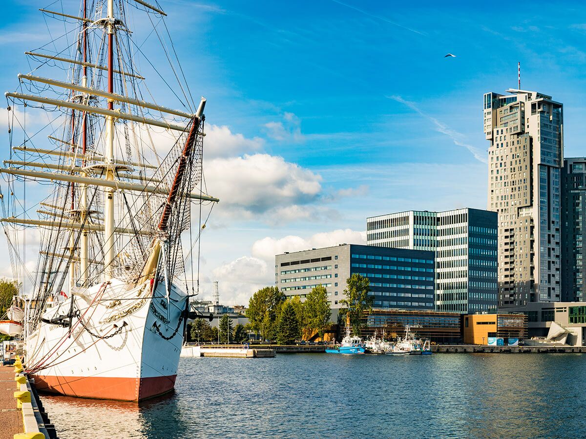 Historisches Segelschiff im Hafen von Gdynia, Polen, vor einer modernen Skyline mit Wolkenkratzern unter blauem Himmel.