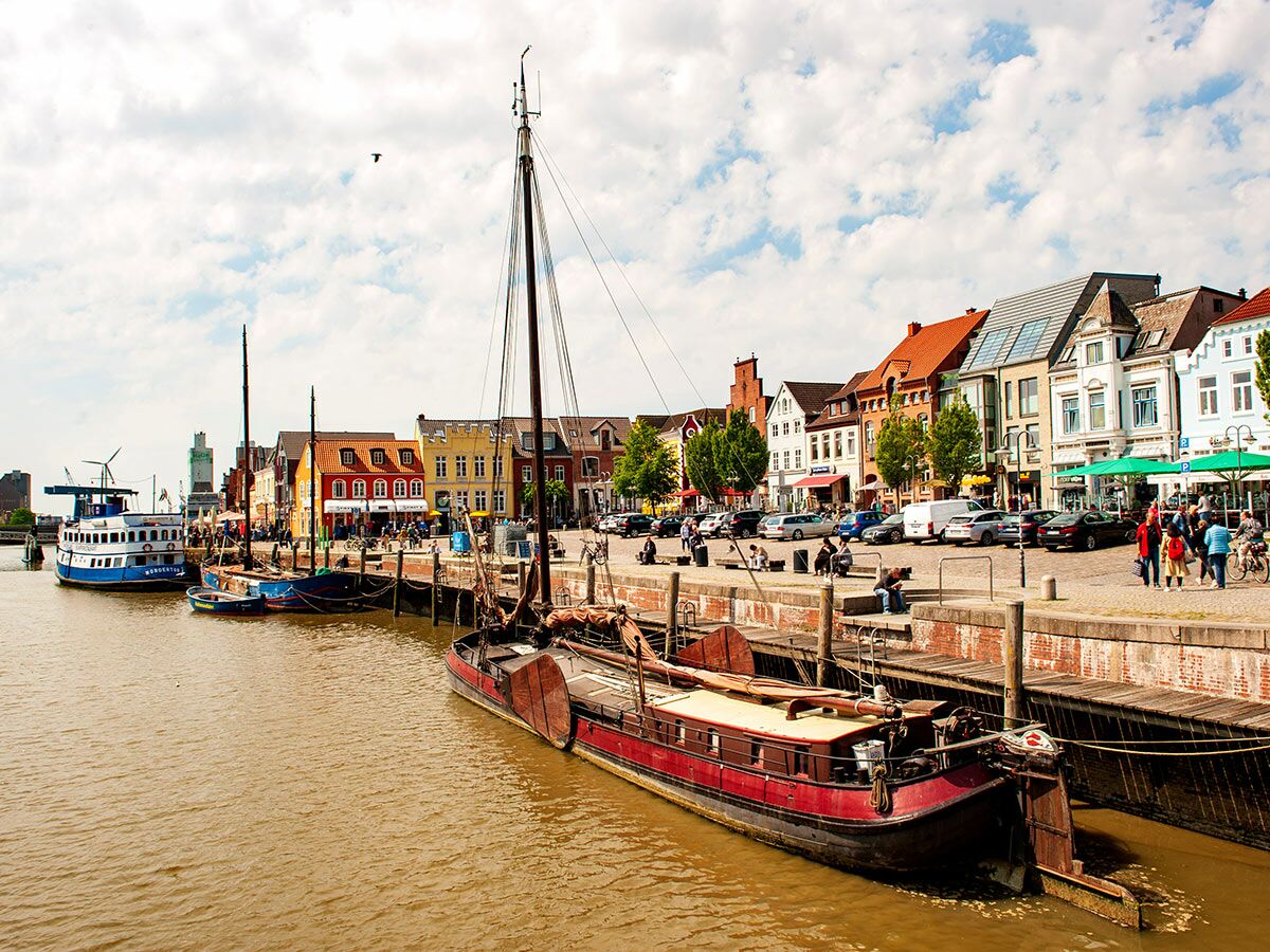 Blick auf eine historische norddeutsche Hafenstadt: bunte Häuser säumen einen Kanal mit Segelbooten, Fähre und Menschen am Kai.