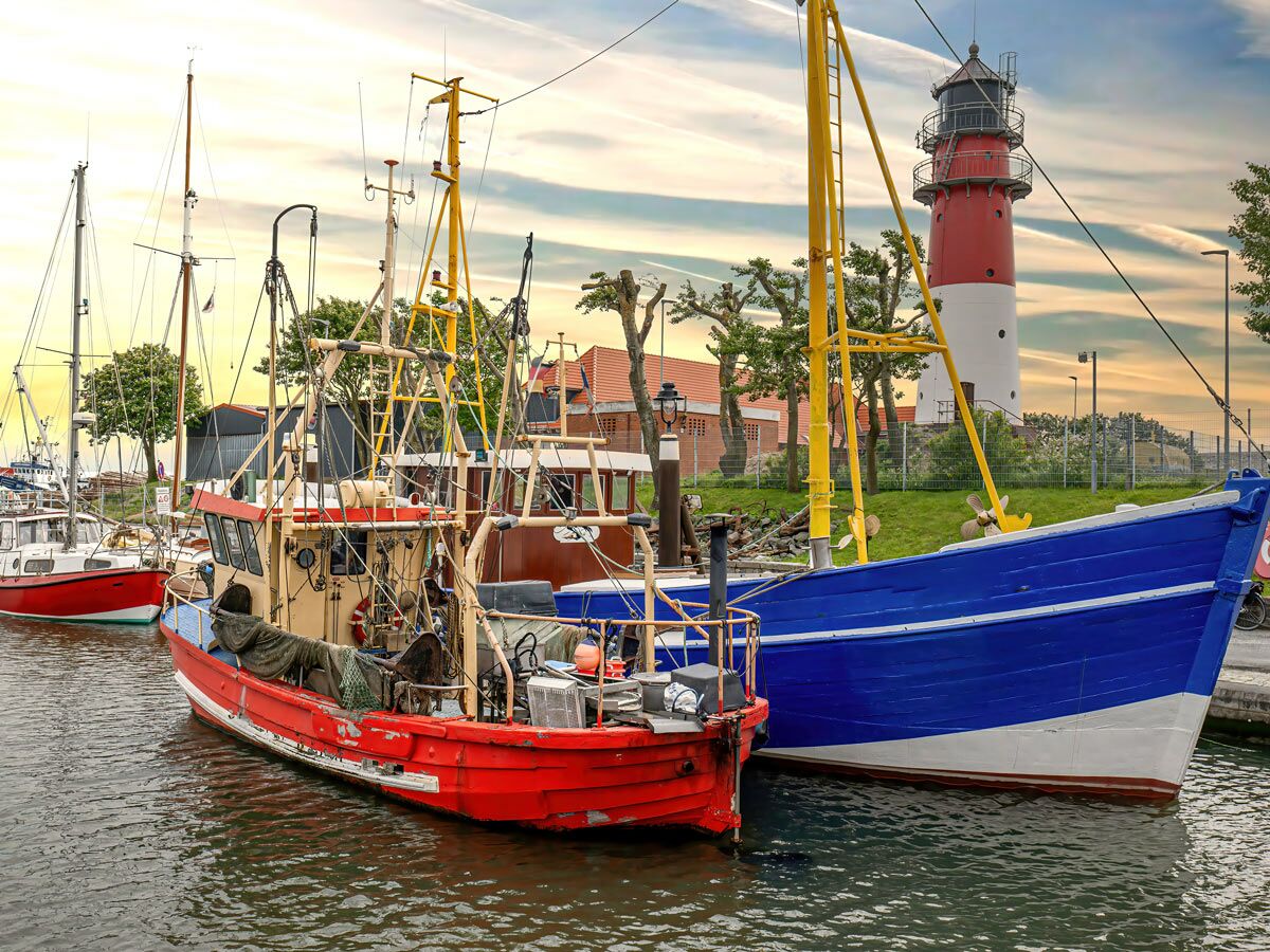 Blick auf einen malerischen Hafen bei Sonnenuntergang mit roten und blauen Fischerbooten im Vordergrund und einem Leuchtturm im Hintergrund.