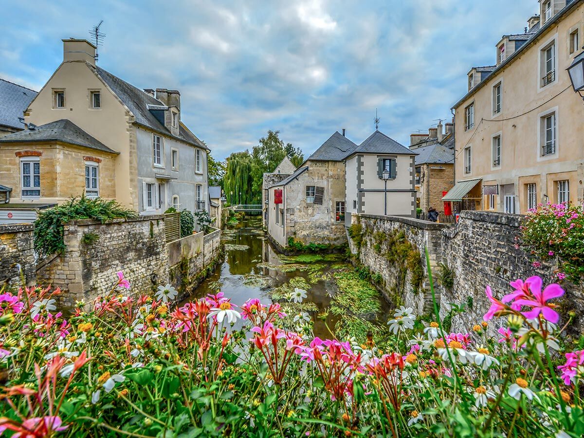 Idyllischer Dorfkanal mit alten Steinhäusern und farbenprächtigen Blumen im Vordergrund unter bewölktem Himmel.