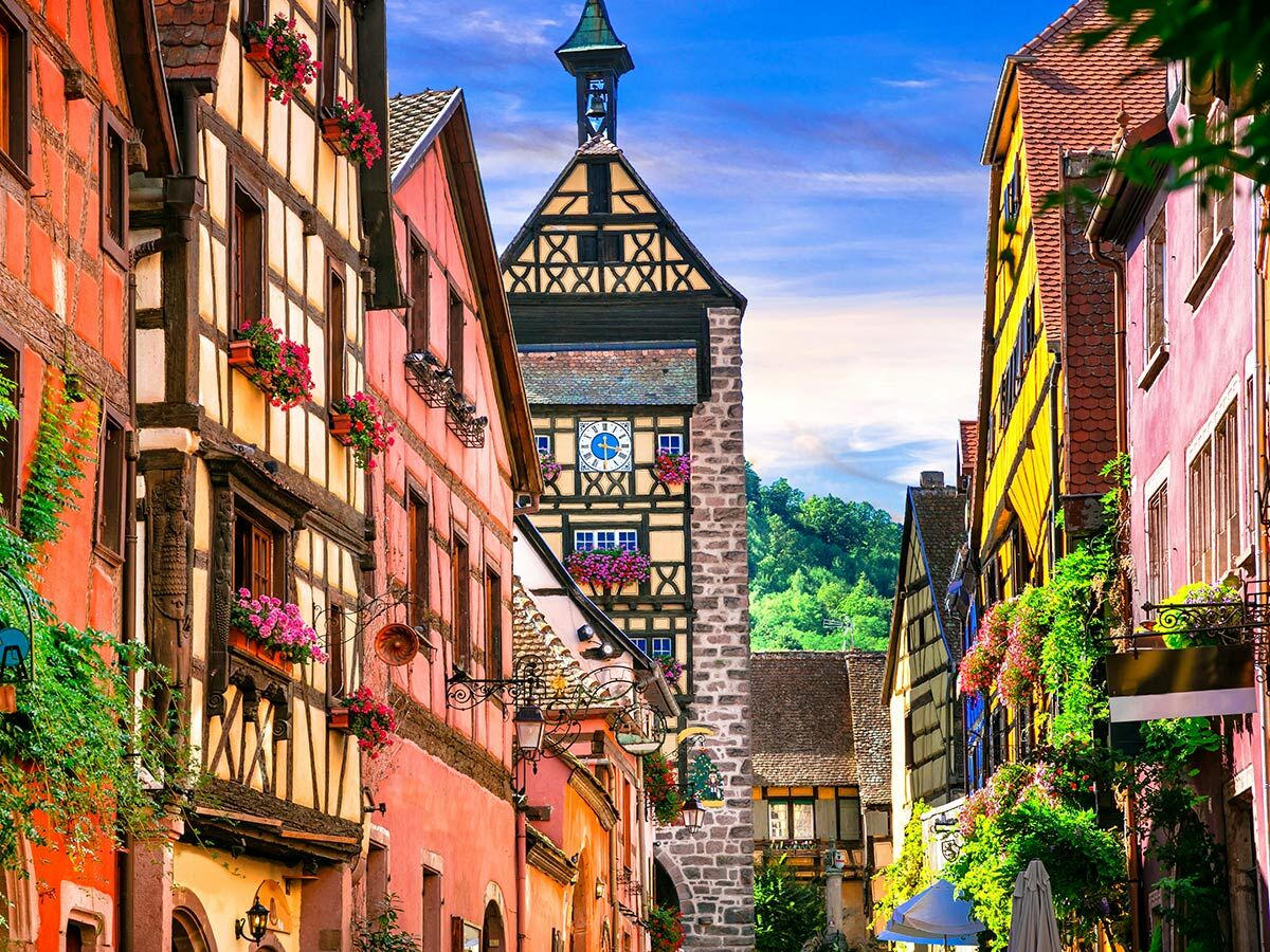 Malerische Altstadtgasse mit farbenfrohen Fachwerkhäusern, blühenden Blumenkästen und einem historischen Uhrenturm im Hintergrund.