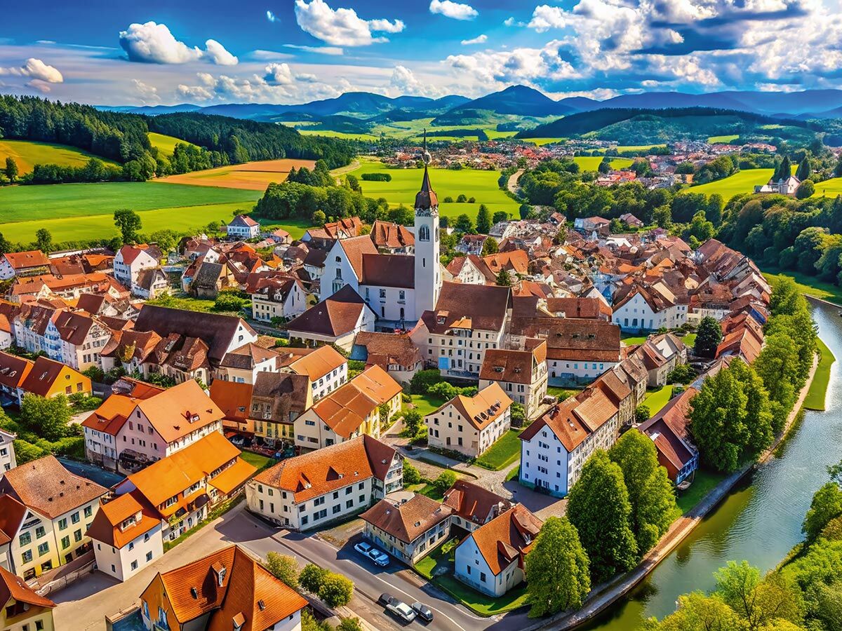 Luftaufnahme eines idyllischen europäischen Dorfes mit rotdachigen Häusern, zentraler Kirche, mäanderndem Fluss und grüner Berglandschaft.