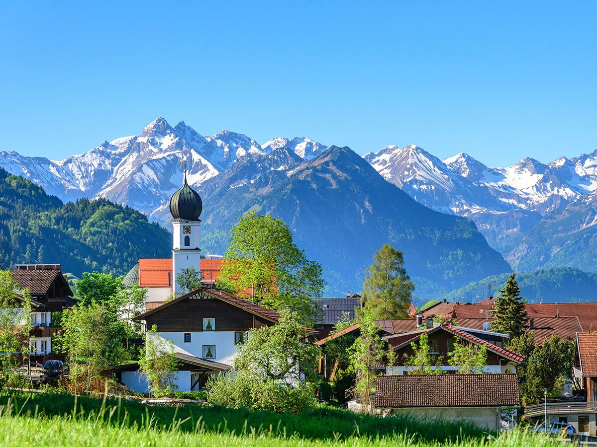 Malerisches Dorf mit weißer Kirche, Zwiebelturm, traditionellen Häusern und schneebedeckten Alpen unter blauem Himmel in Bayern.