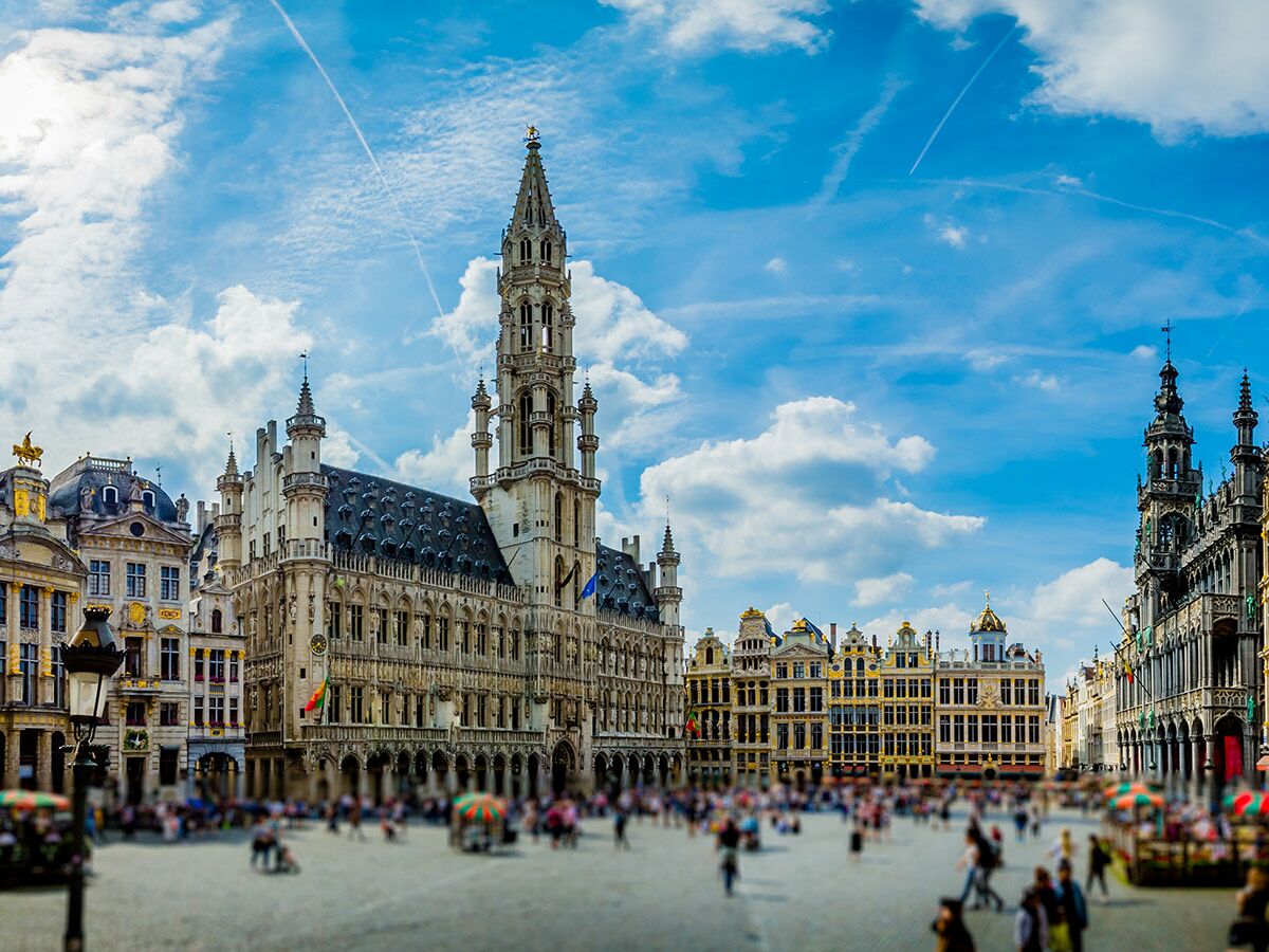 Historischer Grand Place in Brüssel mit dem gotischen Rathaus, den prächtigen Zunfthäusern und vielen Menschen unter blauem Himmel.