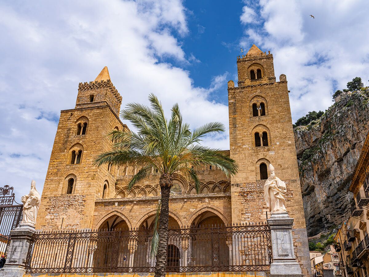 Die Cefalù Kathedrale mit zwei Türmen, Steinmauern und einem Palmenbaum vor felsiger Kulisse in Sizilien, Italien.