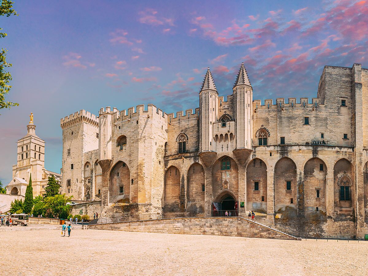 Majestätischer Palais des Papes in Avignon mit Kathedrale und Menschen auf dem Platz unter farbenfrohem Himmel.