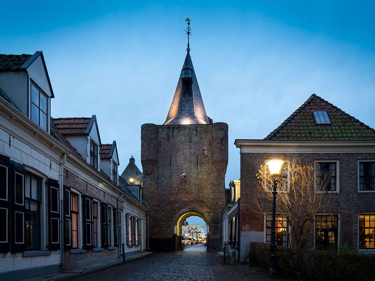 Abendliche Ansicht einer kopfsteingepflasterten Gasse mit altem Backstein-Stadttor, beleuchteten Häusern und Straßenlaterne in Europa.
